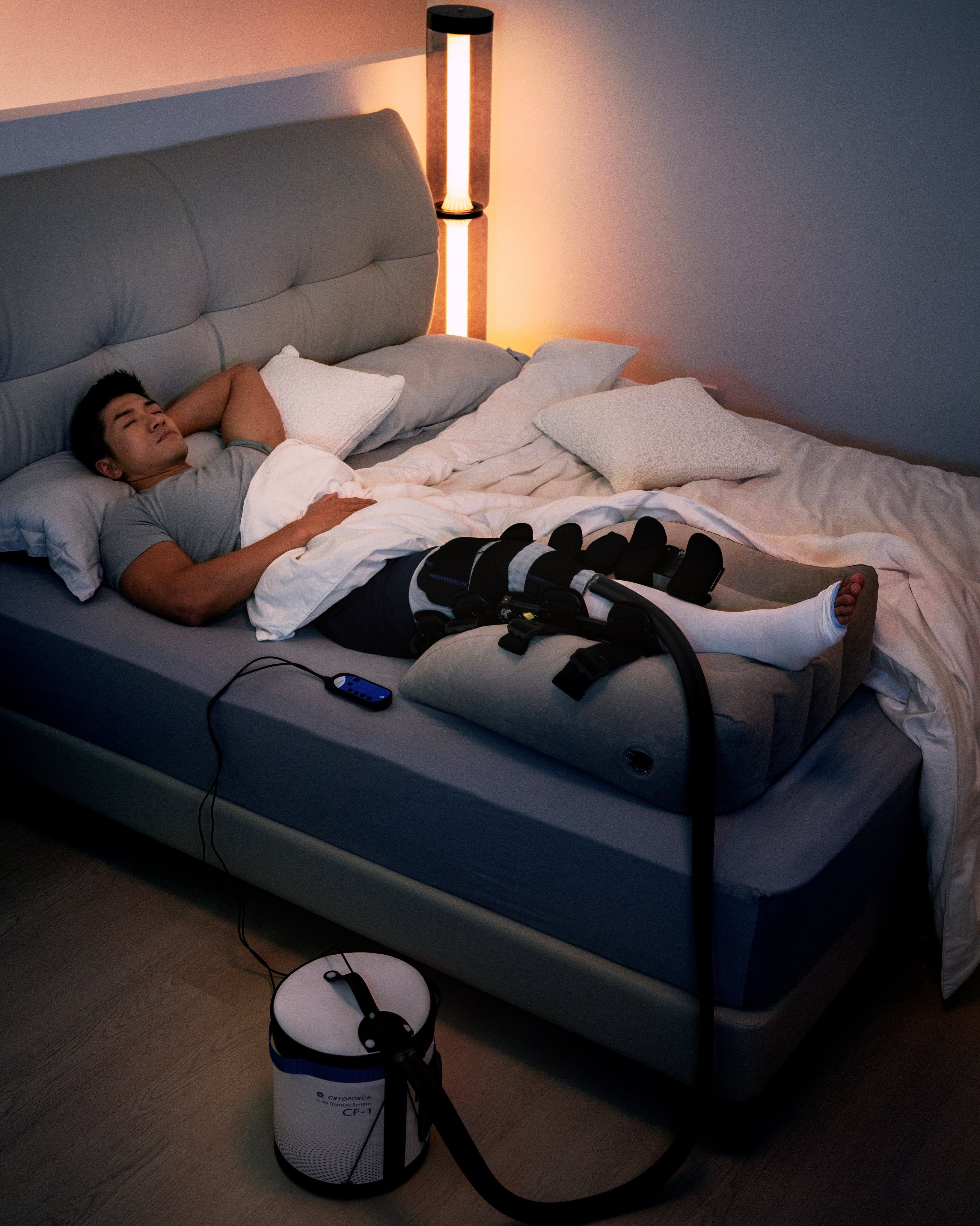 A person lying in bed using a CRYOFORCE cold therapy machine beside the bed in a dimly lit room.