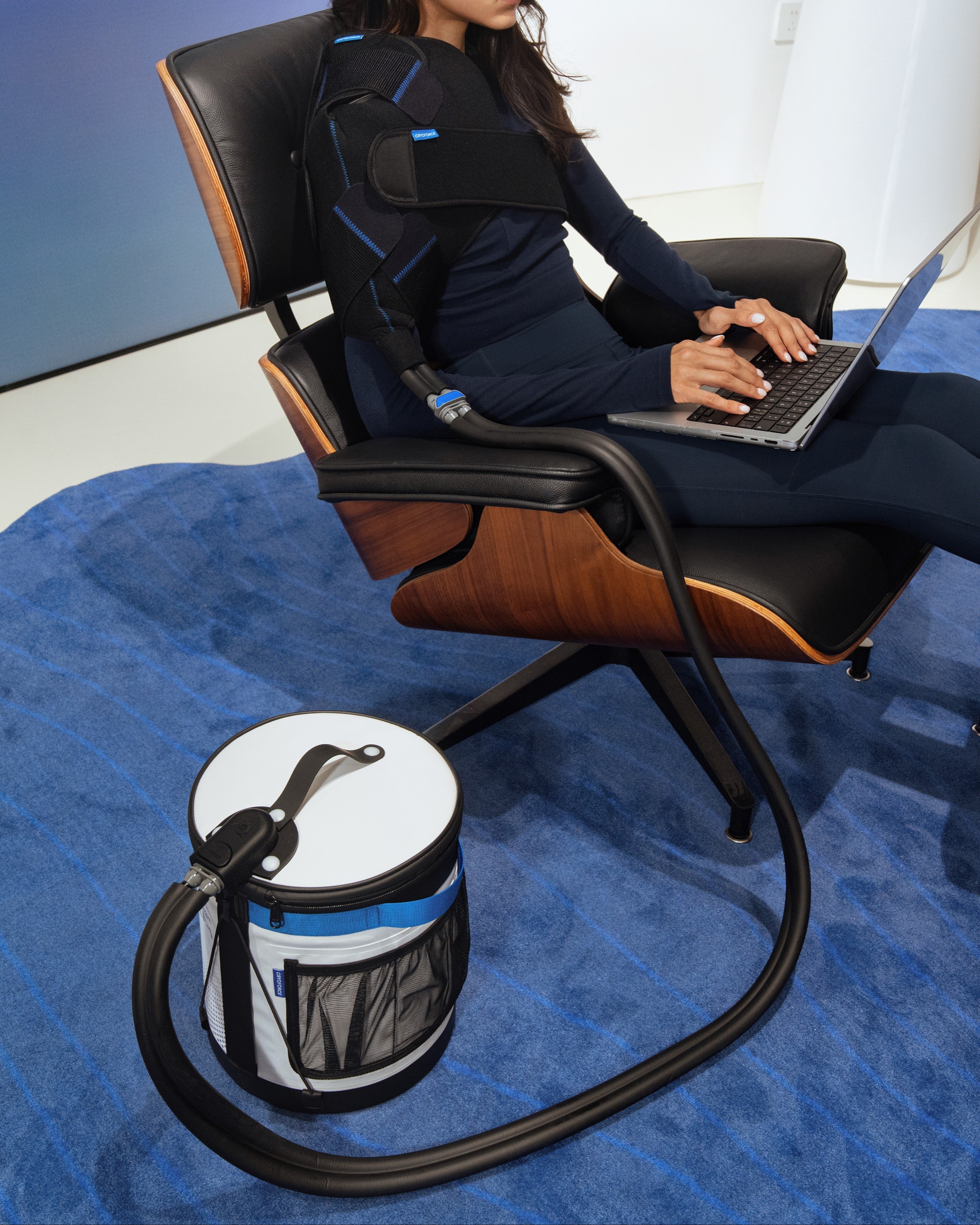 A woman using an ice machine with a black and blue therapy pad on her shoulder, sitting in a modern chair on a blue carpet.